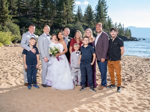 A wedding party poses together on a sandy beach with pine trees and Lake Tahoe's blue waters in the background.