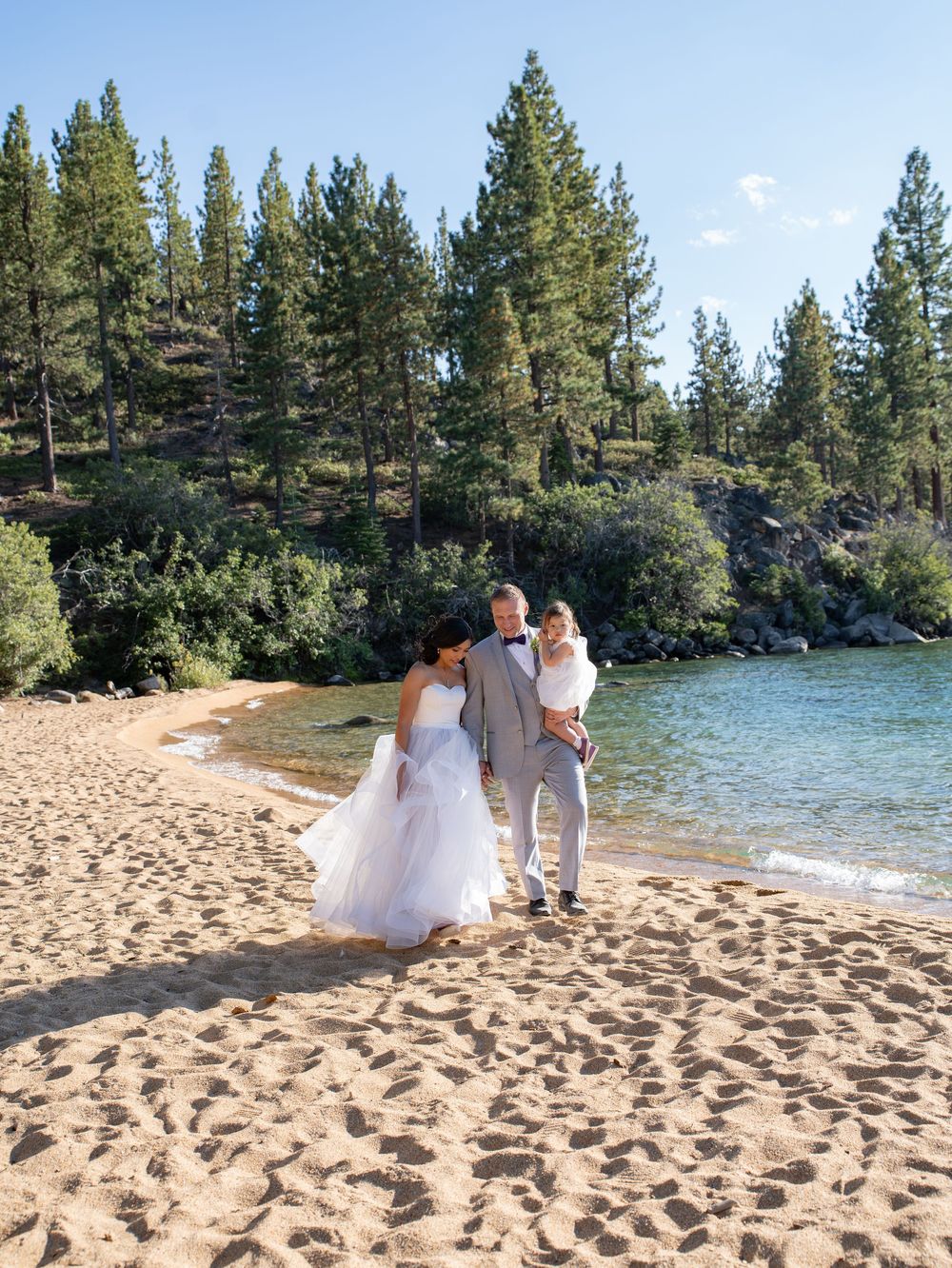 A couple in wedding attire stands together on a sandy beach with evergreen trees and blue lake waters in the background.
