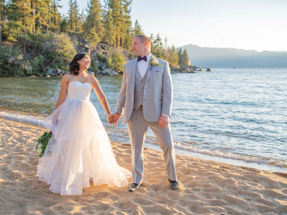Two people walk hand in hand on a sandy beach at sunset with mountains and pine trees in the distance.