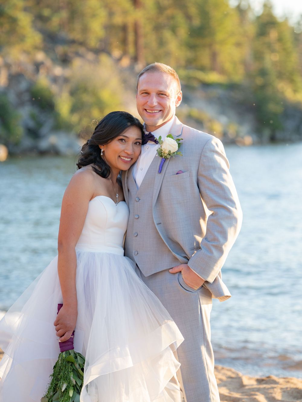 A wedding couple poses together by a lake with pine trees and rocky cliffs in the background on a sunny day.