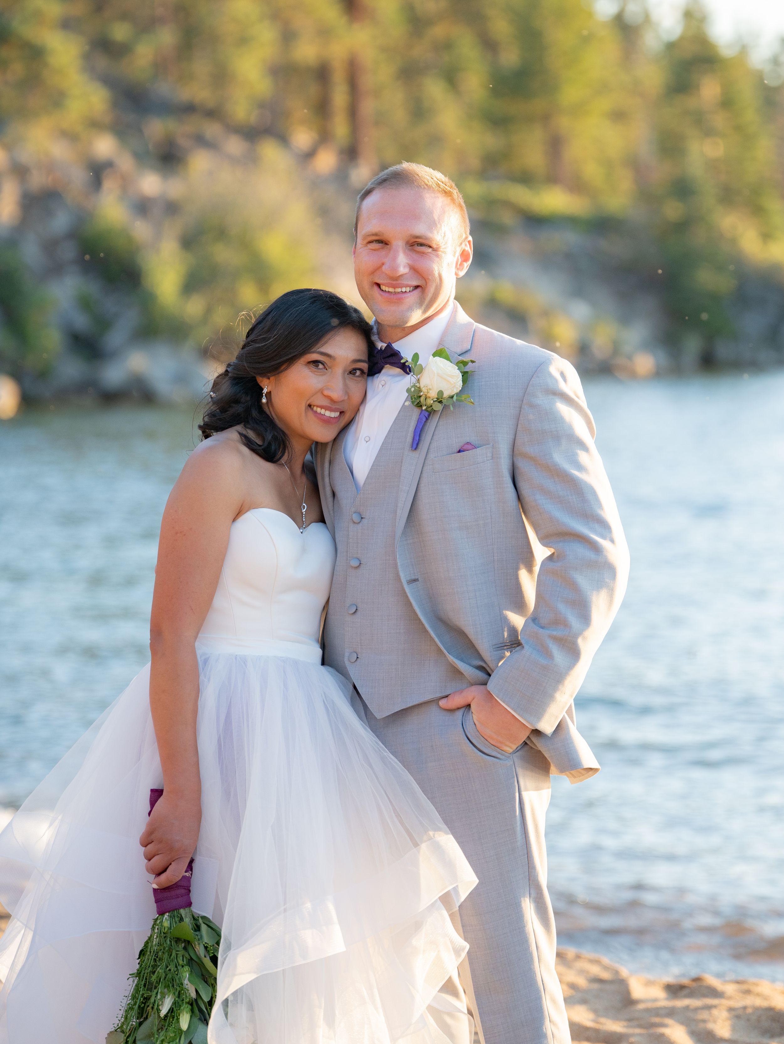 A wedding couple poses together by a lake with pine trees and rocky cliffs in the background on a sunny day.