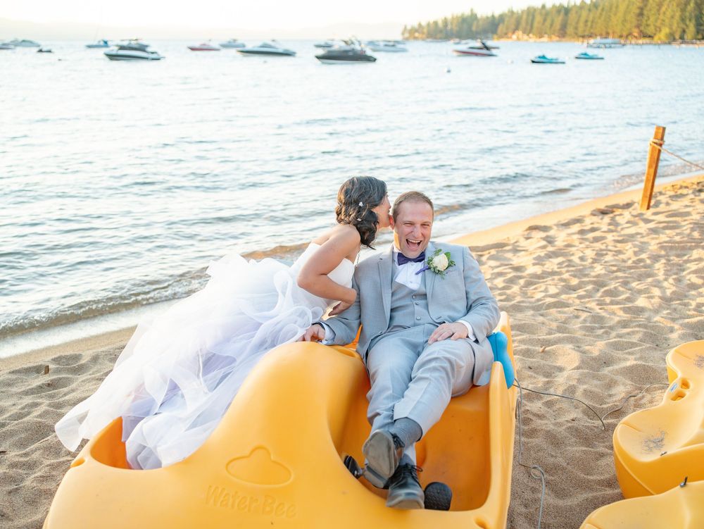 A couple shares a romantic moment in yellow pedal boats at the beach with boats and trees in the background.