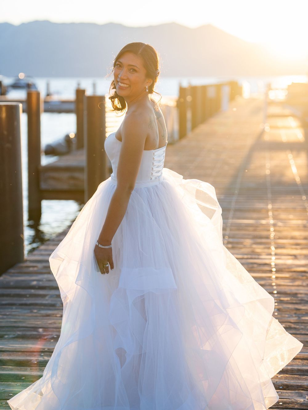 A bride in a flowing white dress twirls on a wooden dock at sunset with mountains and water in the background.