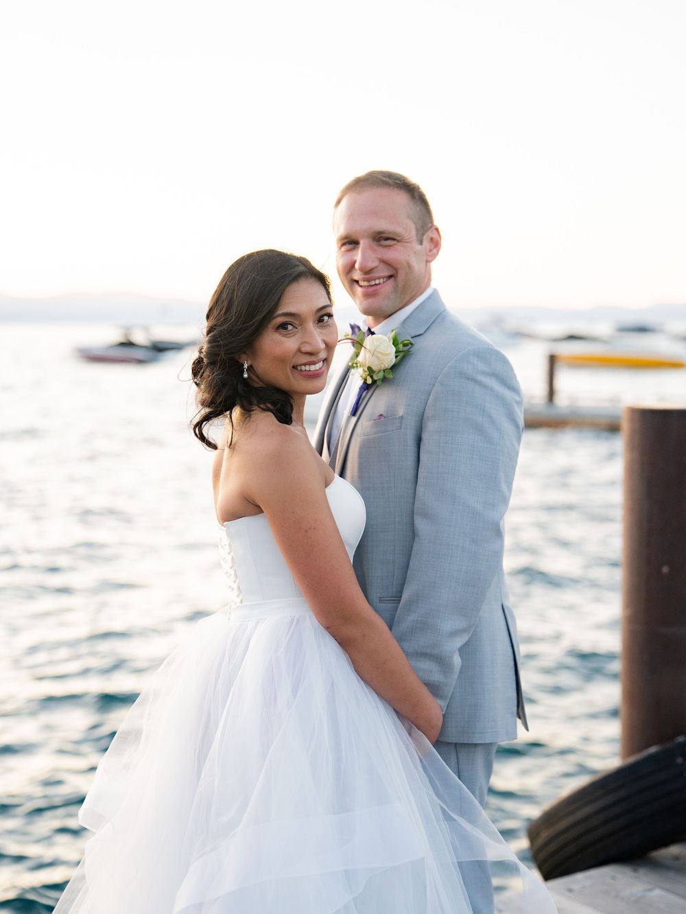 A bride and groom share a romantic moment by the waterfront with boats in the background at sunset.