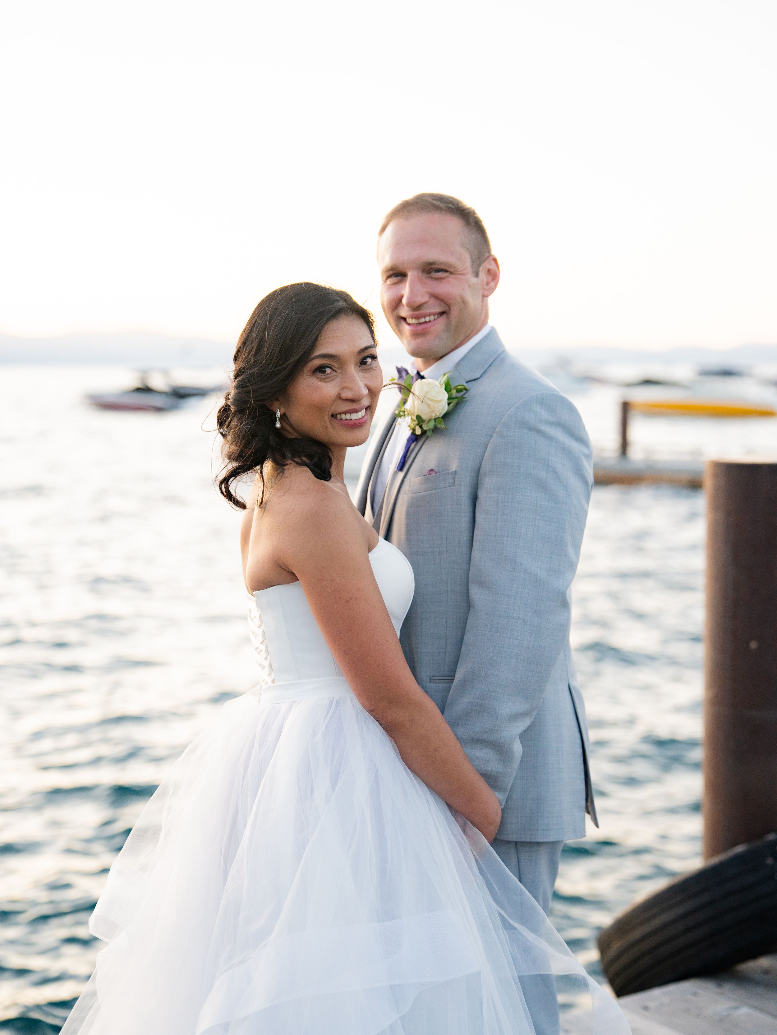 A bride and groom share a romantic moment by the waterfront with boats in the background at sunset.
