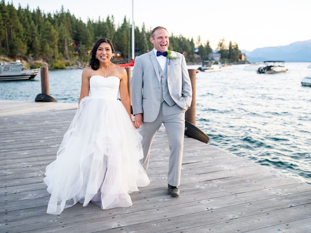 A couple walks hand in hand on a wooden dock by a lake surrounded by evergreen trees and mountains.