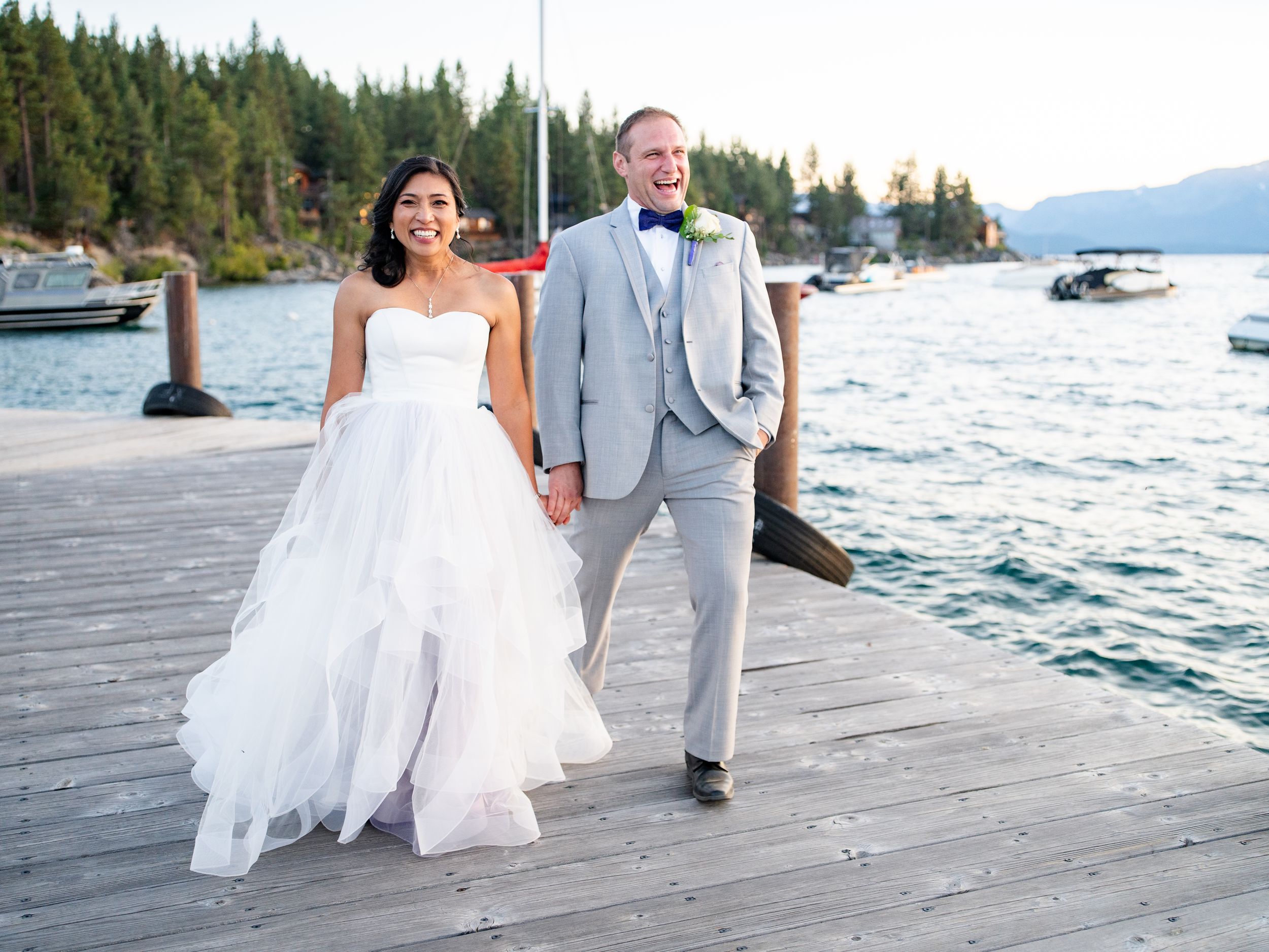 A couple walks hand in hand on a wooden dock by a lake surrounded by evergreen trees and mountains.