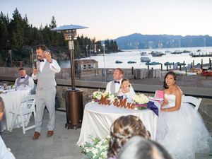 An outdoor wedding reception on a lakeside deck at dusk with mountains in the background and white floral decorations.