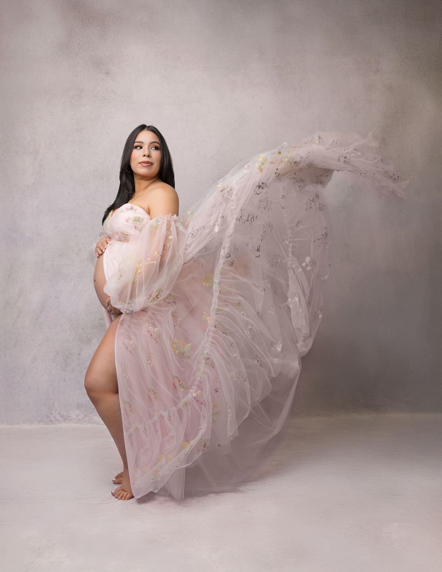 A maternity photoshoot features a flowing pink tulle gown creating ethereal movement against a gray studio backdrop.