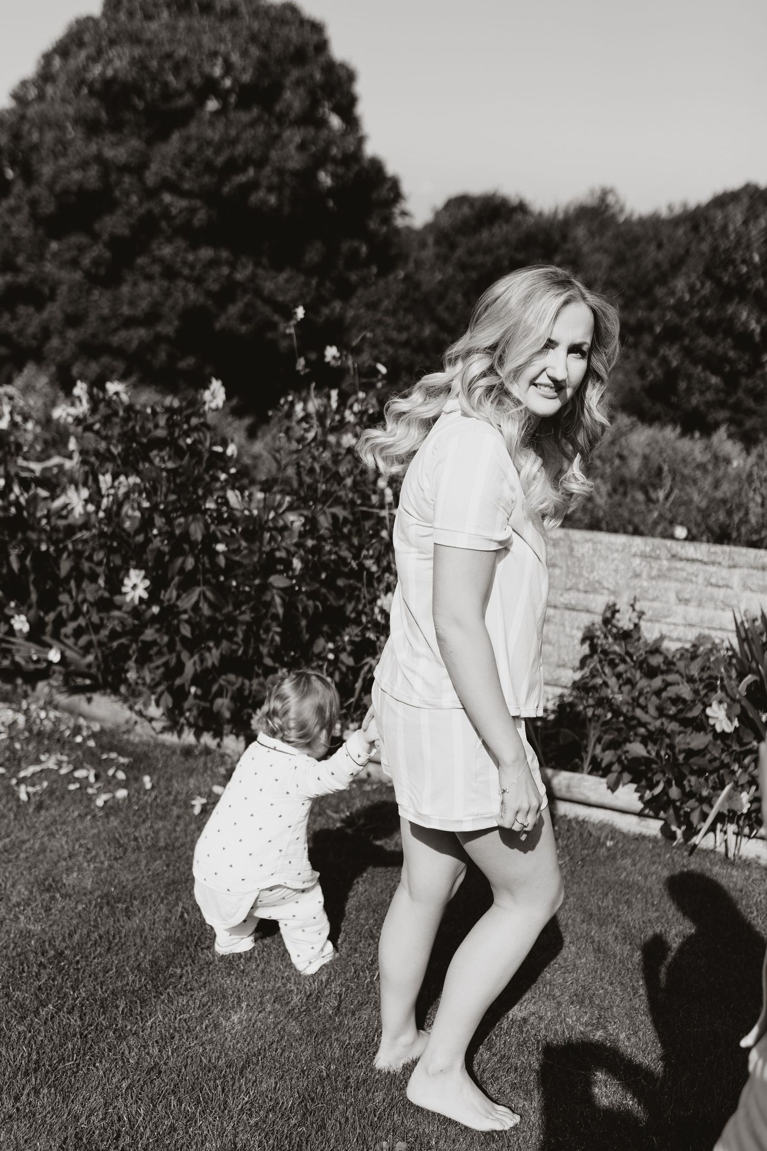 Black and white photo of mother and toddler walking in a garden on a sunny day.