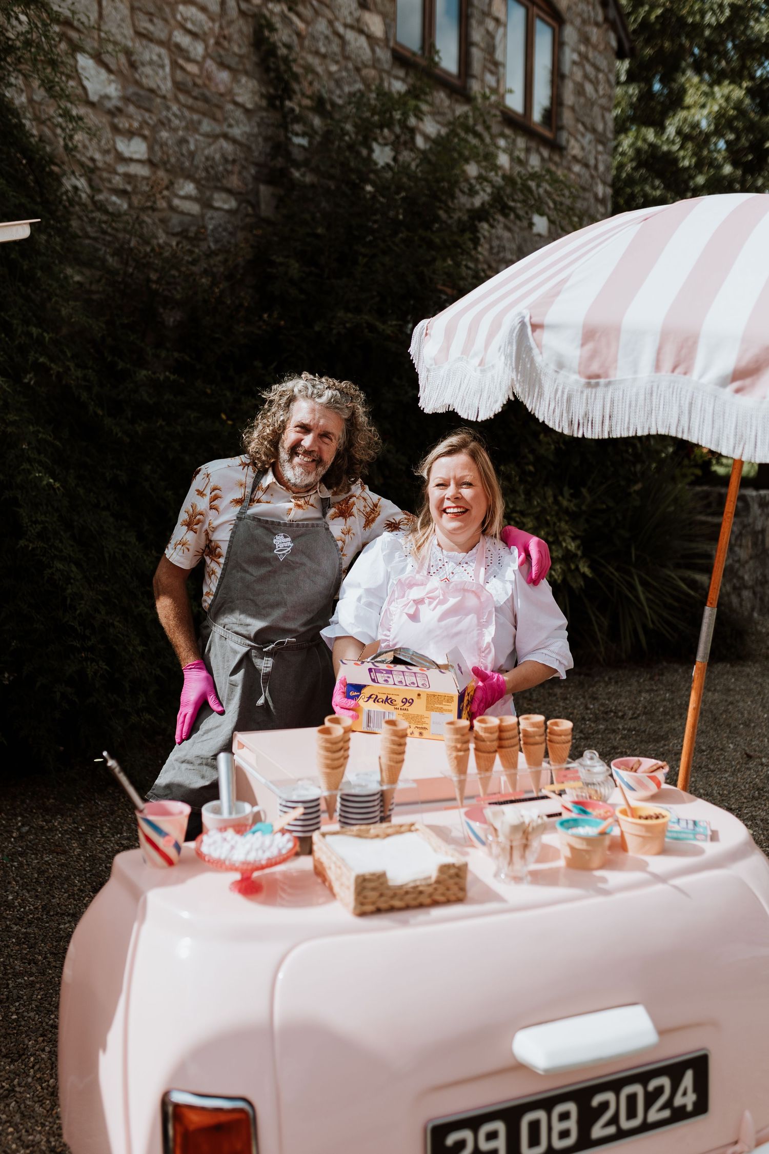 Ice cream vendors serve customers at an outdoor dessert stand with parasols and cones displayed on a pink tablecloth.