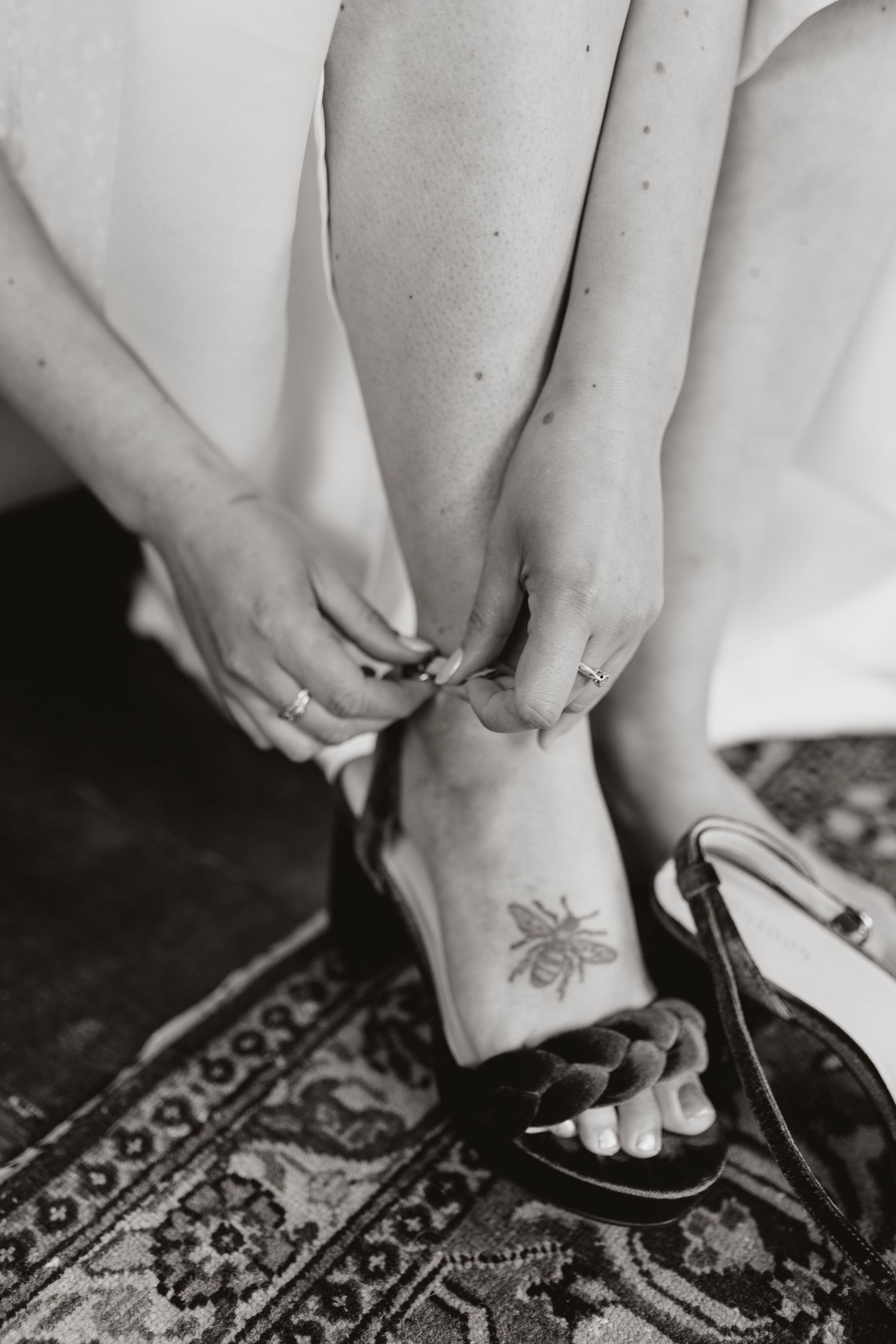 Black and white photograph of hands adjusting a heel strap with a small bee tattoo visible on the foot.