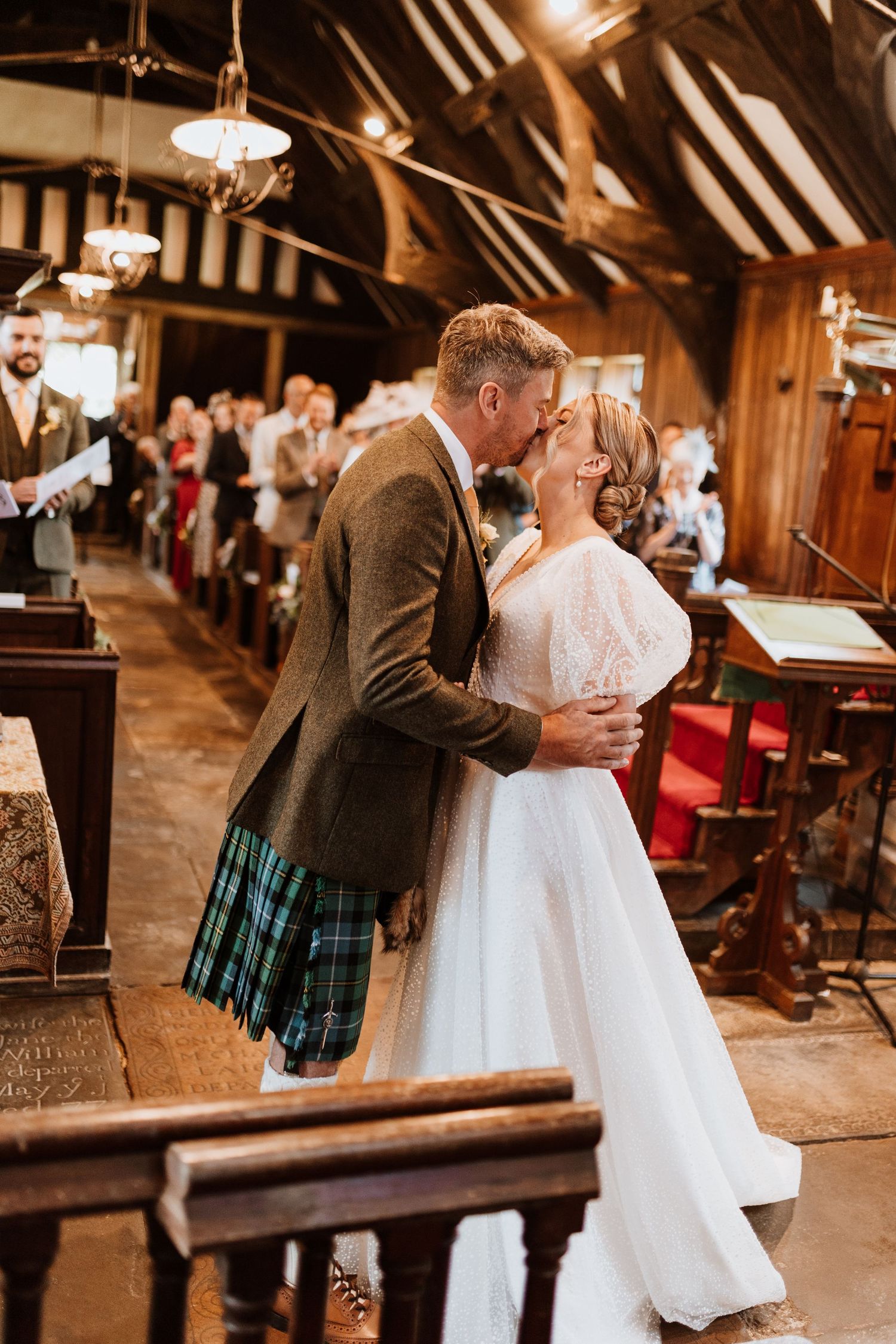 A romantic moment captured in a rustic barn venue featuring traditional Scottish attire and a white wedding dress.