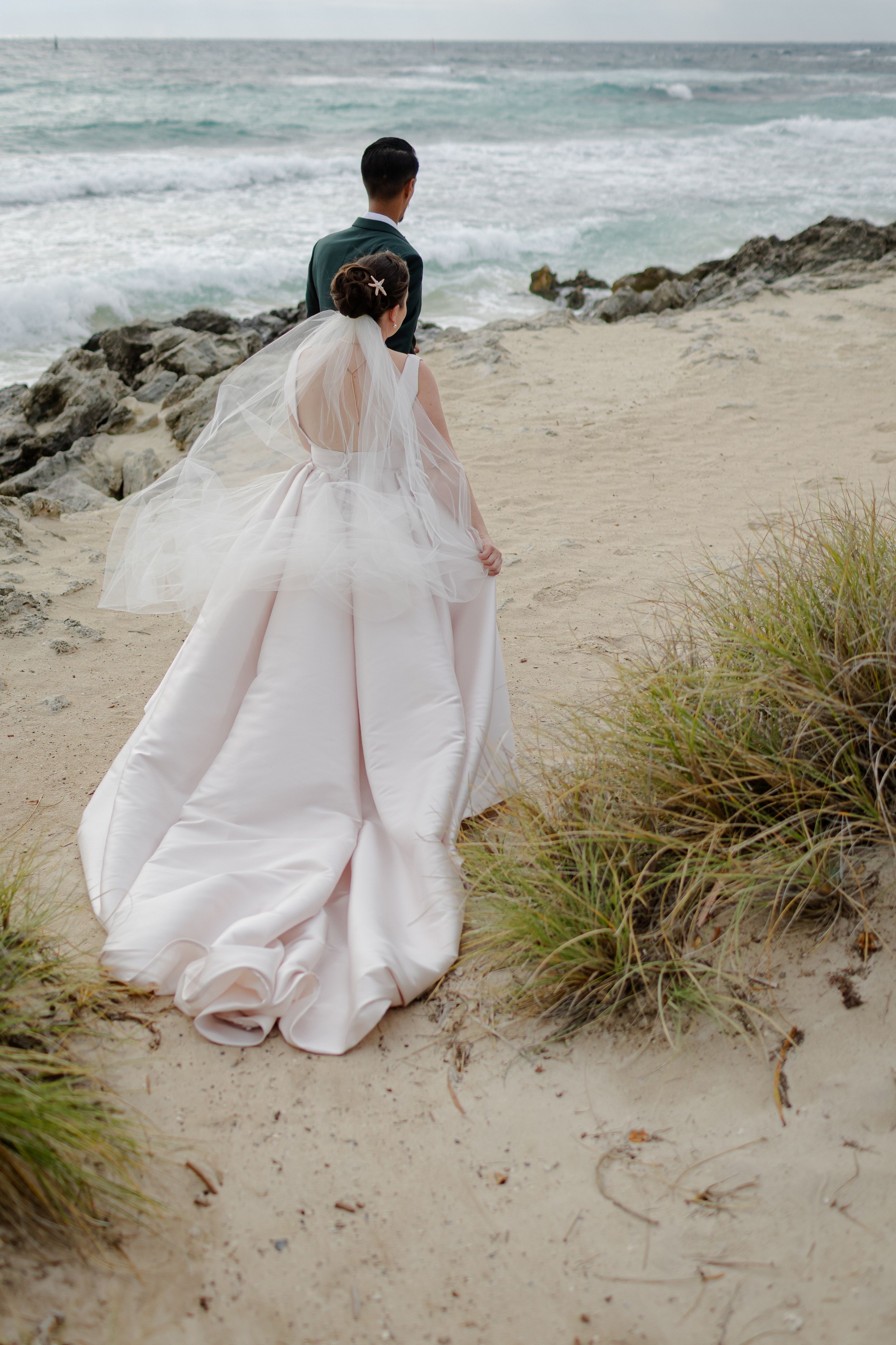Person wearing a flowy white wedding dress sitting on beach sand near ocean with coastal vegetation against dramatic sky.