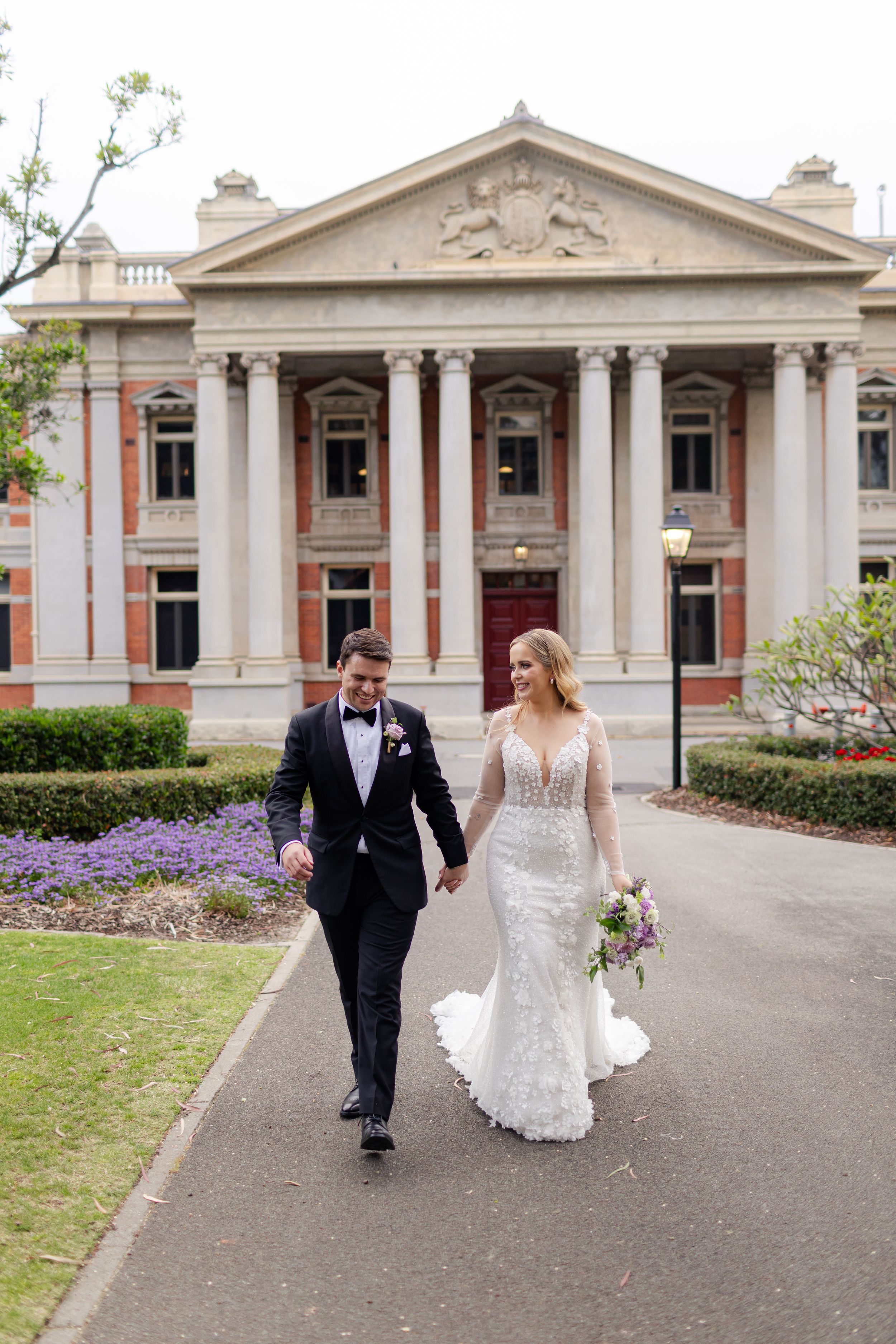 A couple in wedding attire walking together on a path in front of a grand historic building with classical columns.