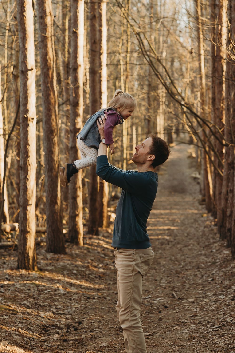 An adult lifts a small child in the air while standing on a tree-lined path in a sunlit autumn forest.