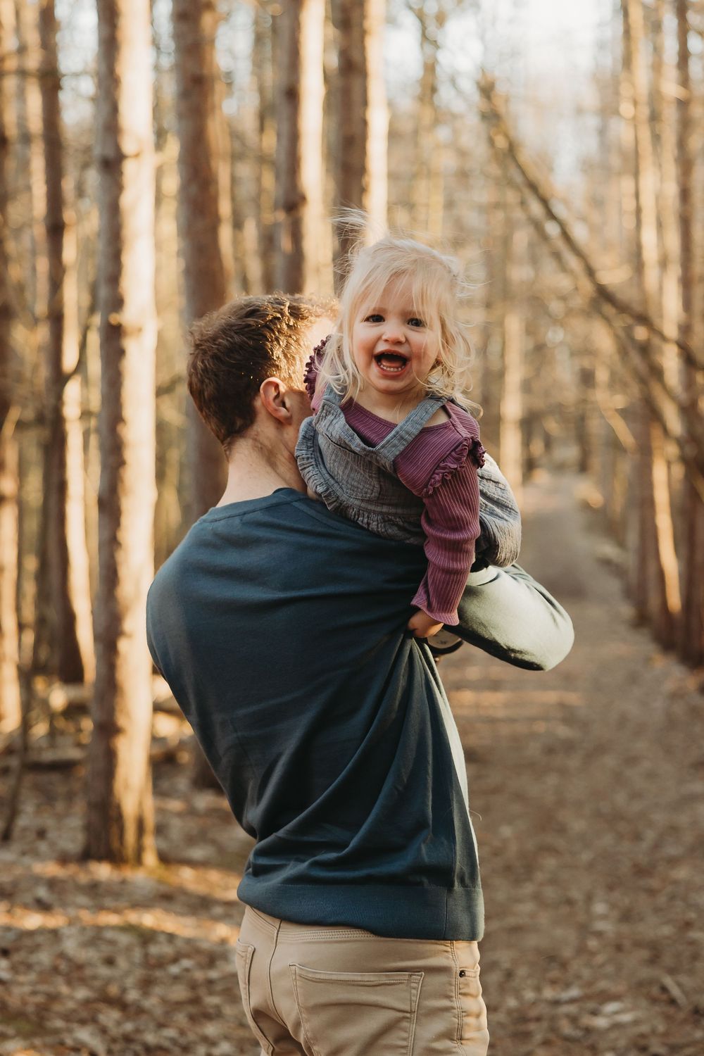 Someone in a navy sweatshirt holds a toddler while walking through a sunlit autumn forest.
