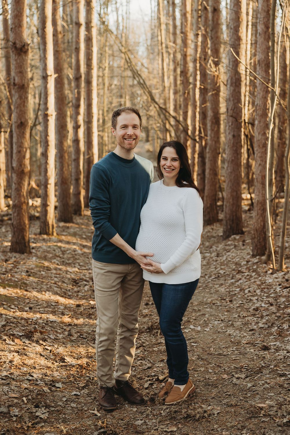An expectant couple stands together on a forest path surrounded by tall trees in warm golden light.