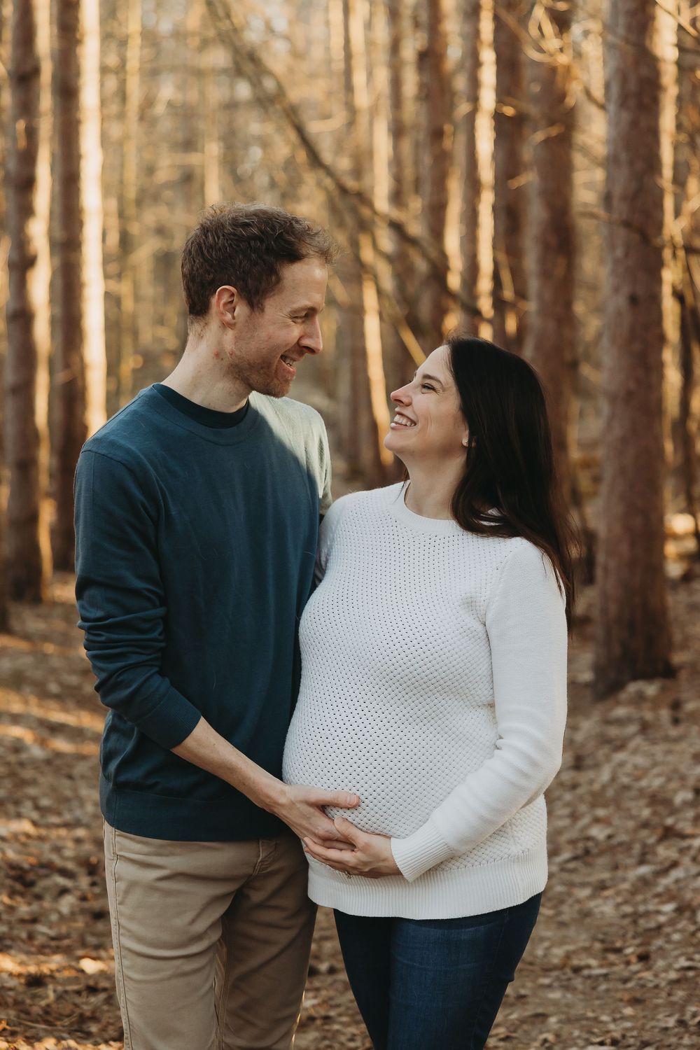 A couple shares an intimate moment while embracing in an autumn forest setting as they await their new arrival.