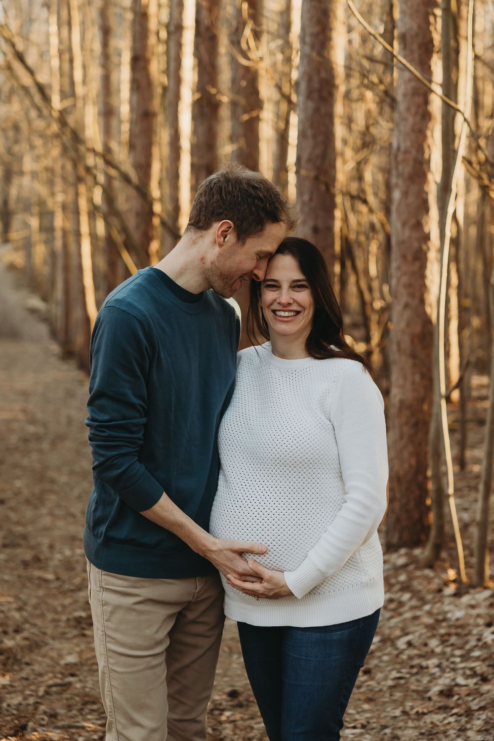 A couple in casual sweaters embrace while standing among tall trees in an autumn forest at sunset.