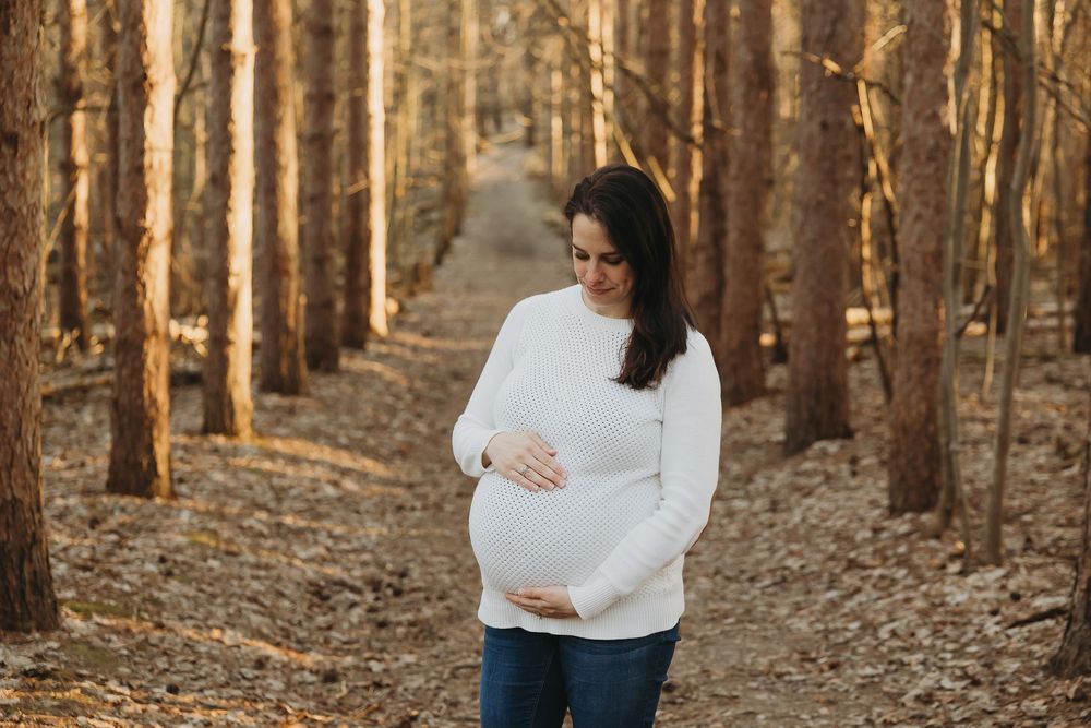 A person in a white sweater holds their baby bump while standing in a sunlit forest during an autumn maternity photoshoot.