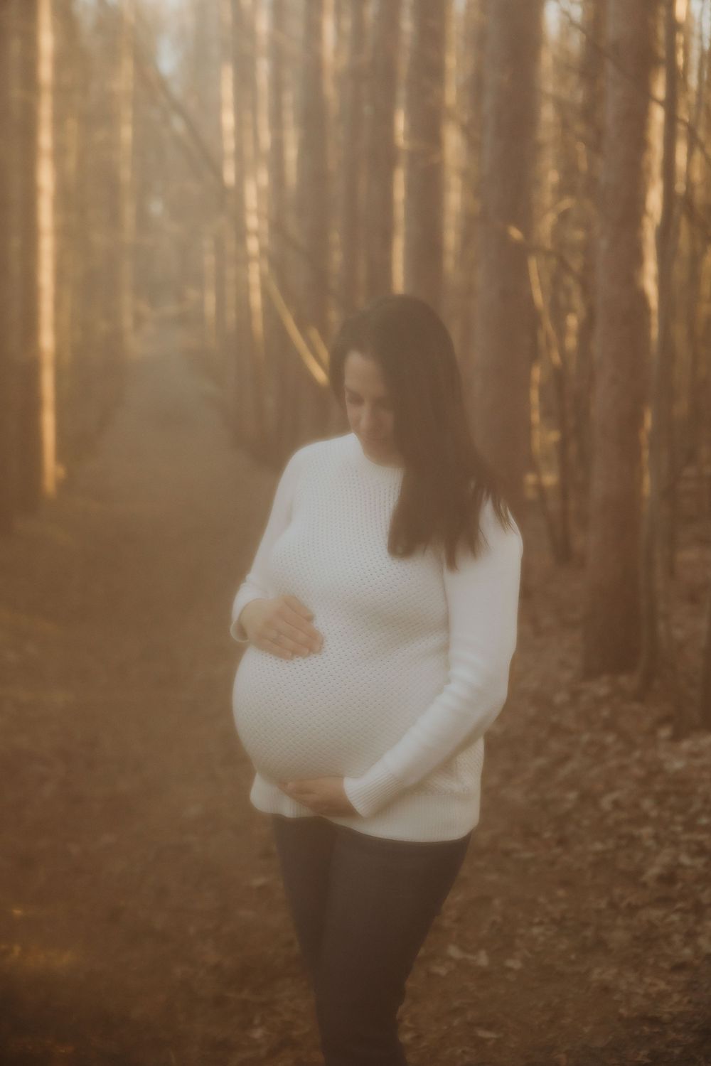 A person in a white sweater stands in a sunlit forest while cradling their pregnant belly.
