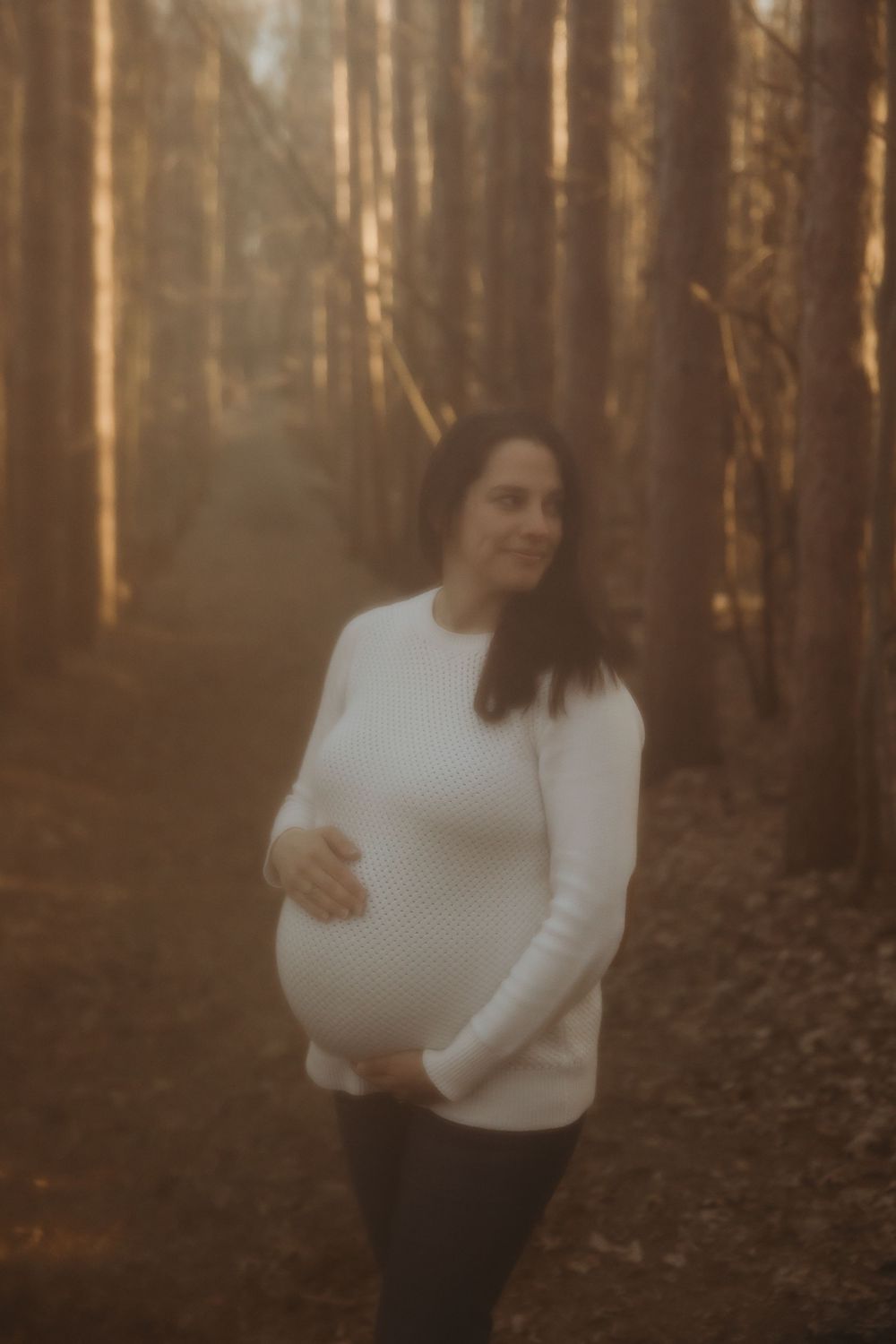 A woman in a white sweater stands among tall trees in a moody autumn forest during golden hour.