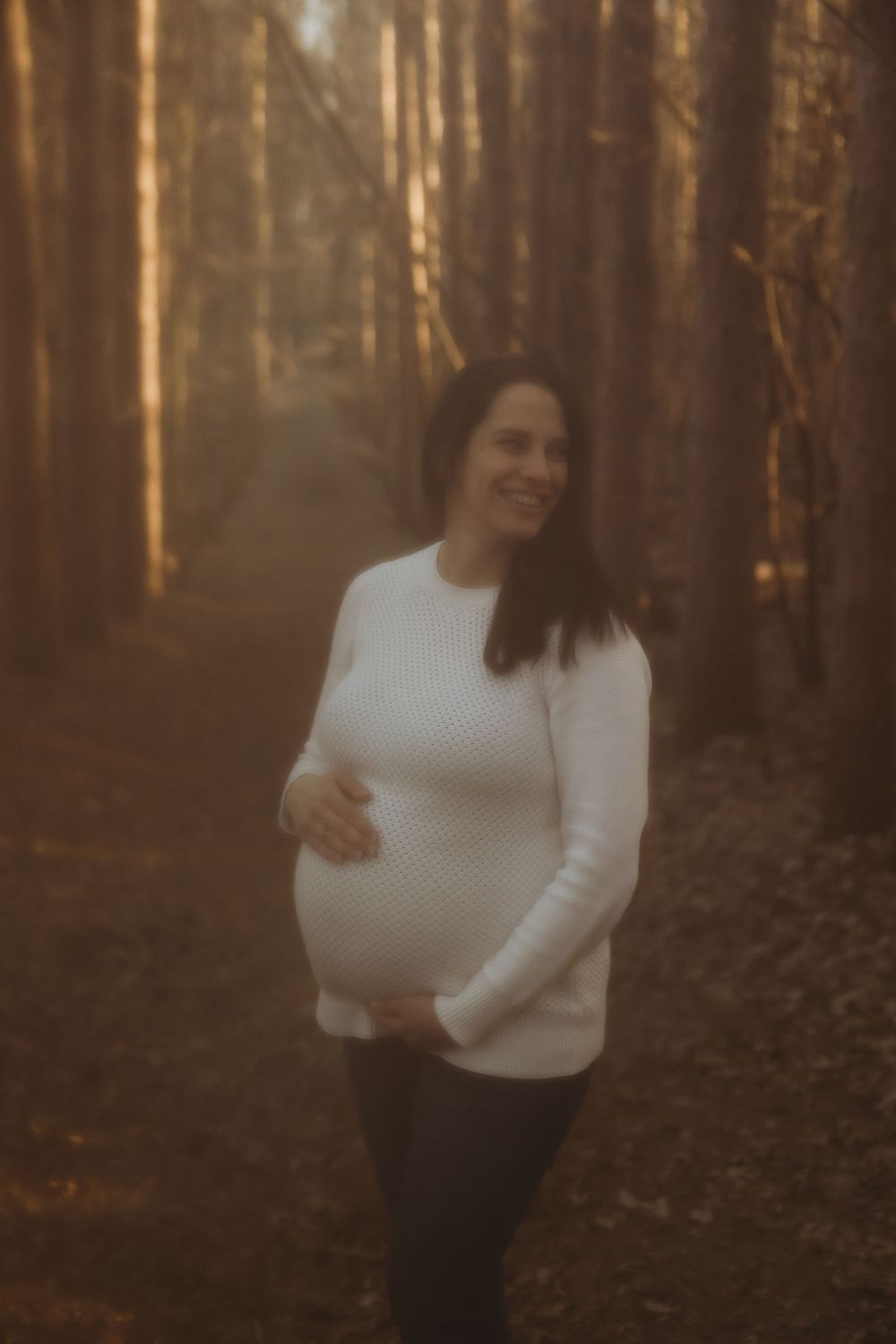 A woman in a white sweater stands in a sunlit autumn forest with golden evening light filtering through trees.