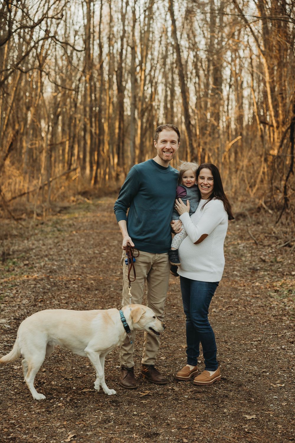 A couple stands together on a forest trail with their yellow Labrador retriever on an autumn afternoon.