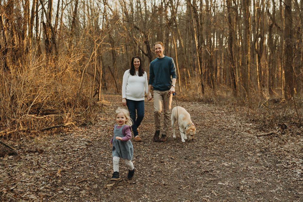 Two people walking together with their dog on a fallen leaves covered trail through bare autumn woods.