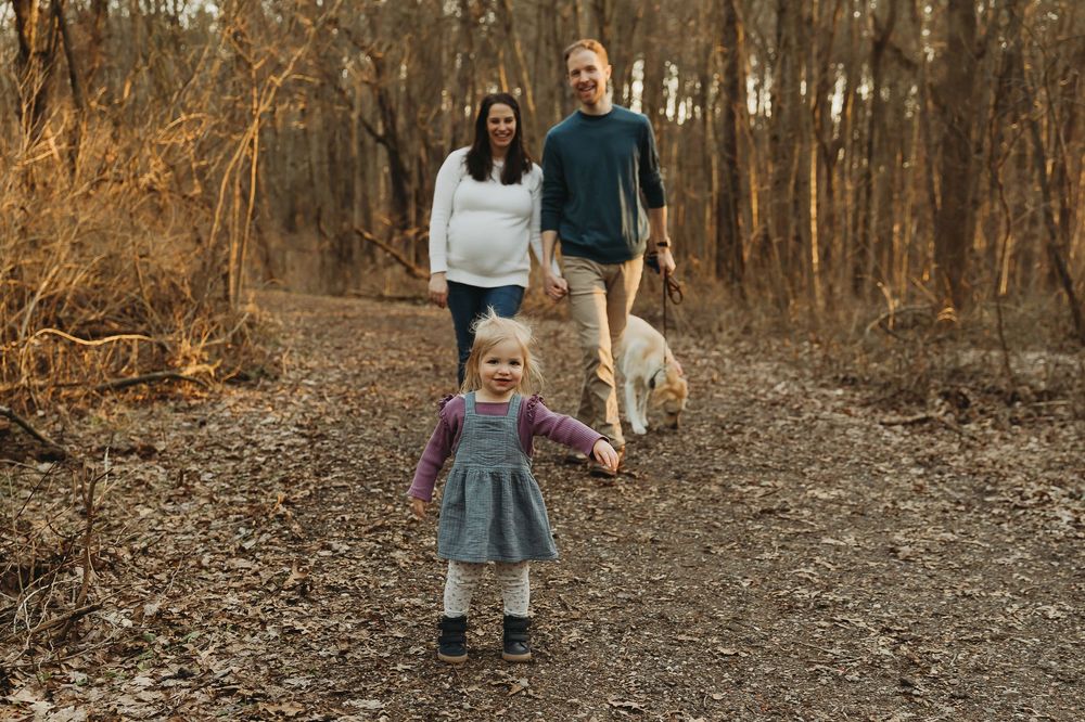 A family and their dog walk together on an autumn trail through a wooded area with golden brown foliage.