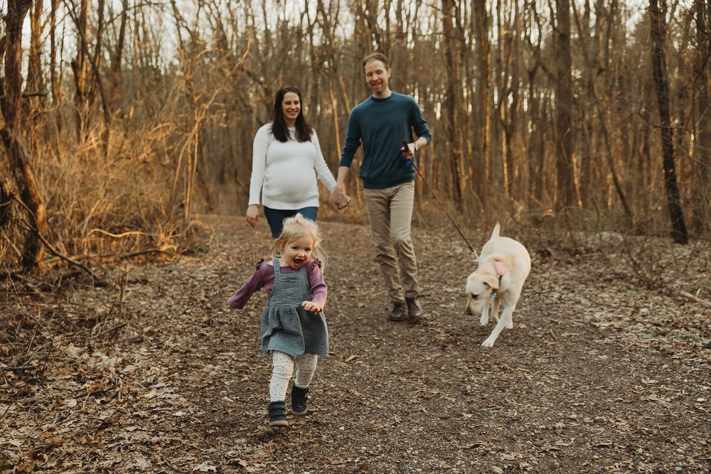 A family and their white dog enjoy an autumn walk together on a wooded trail covered in fallen leaves.