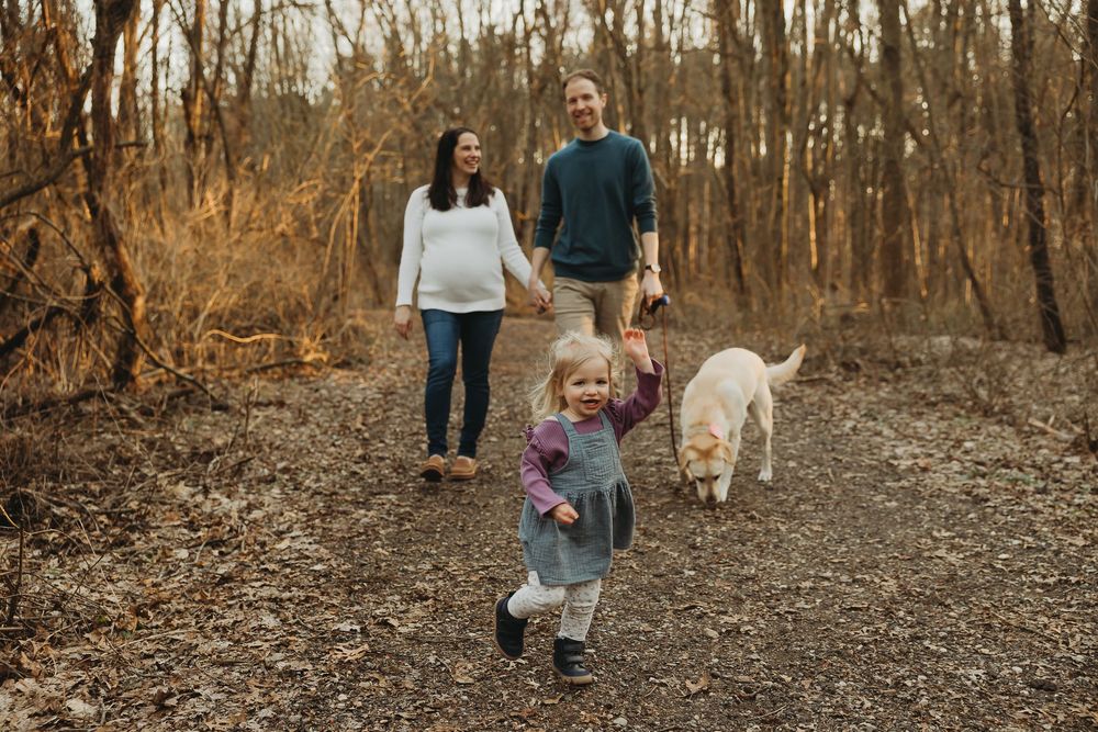 A toddler runs ahead on a forest path while walking with parents and a white dog on an autumn afternoon.