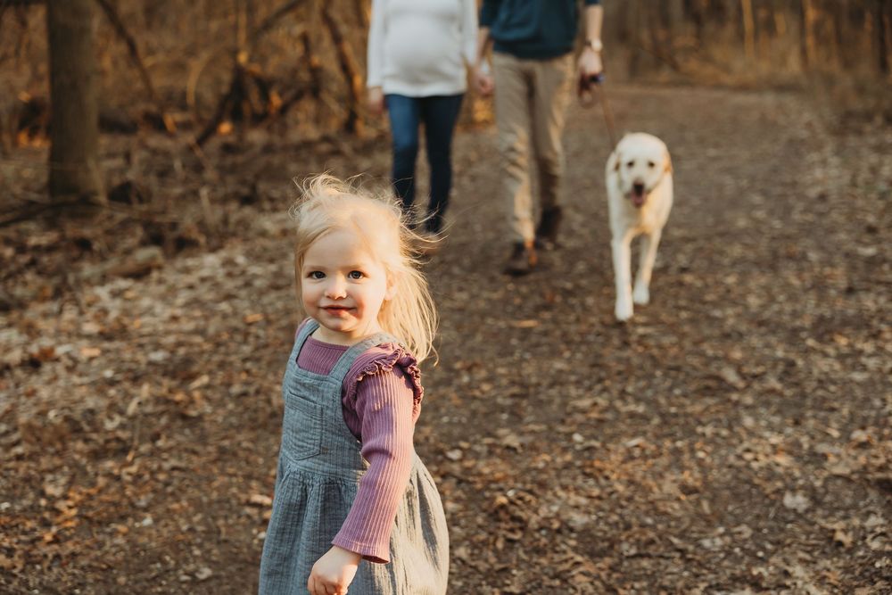 Young toddler wearing a pink sweater explores a wooded trail with family members on an autumn afternoon.