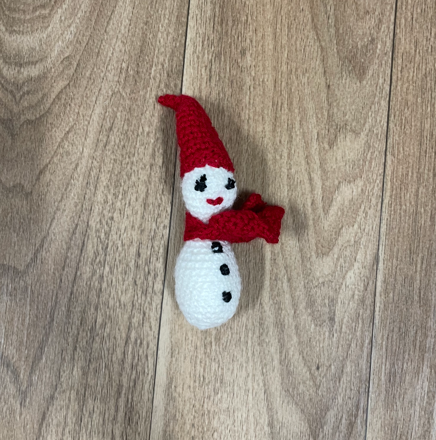 A small crocheted snowman wearing a red hat and scarf on wooden floor.