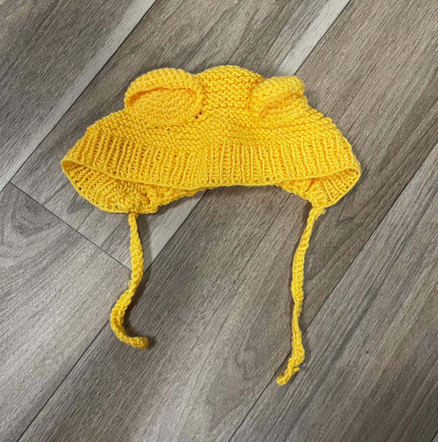 A bright yellow knitted baby bonnet with ties lying on a wooden floor.
