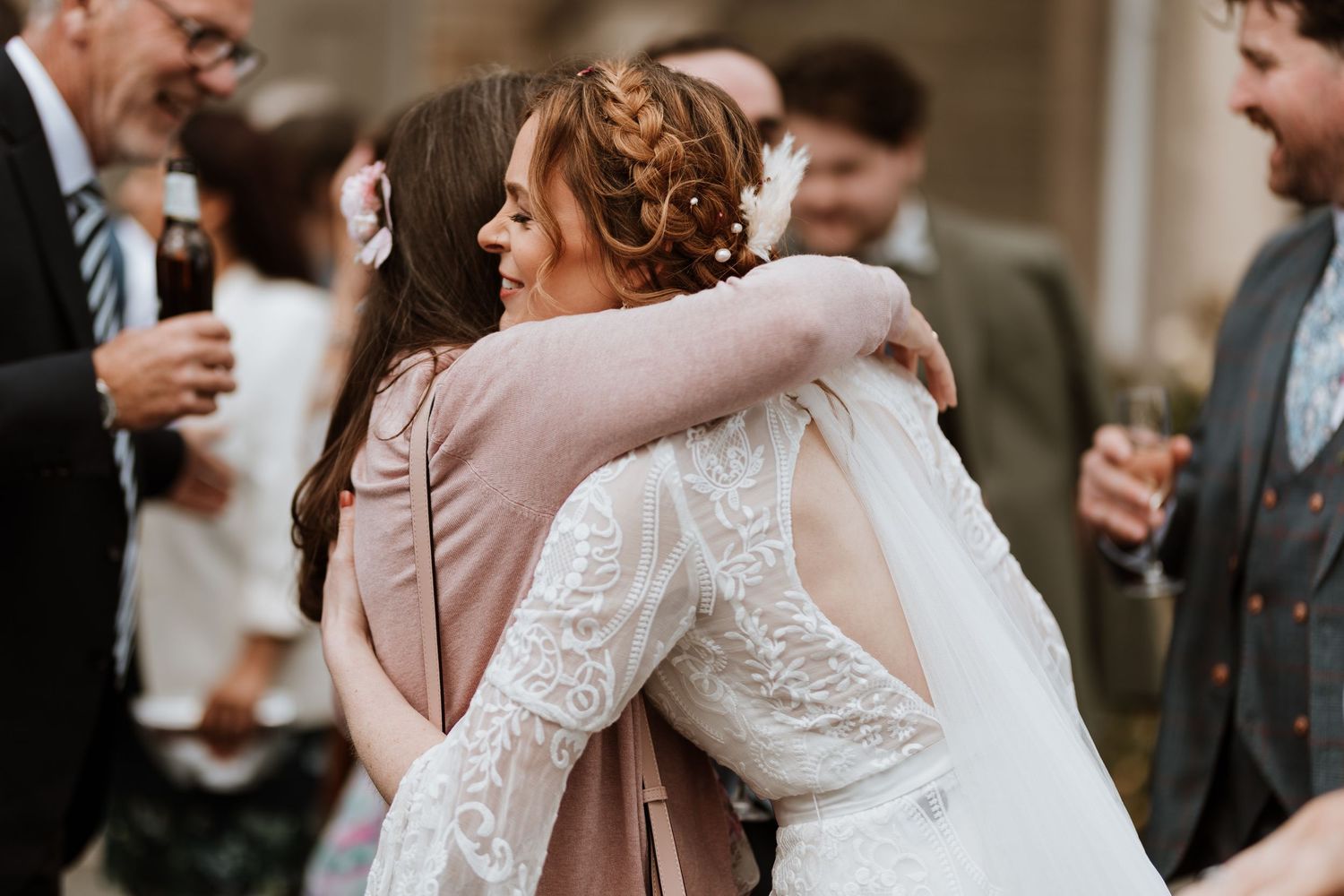 A person in a beautiful lace wedding dress hugs another guest wearing pink at a wedding reception.