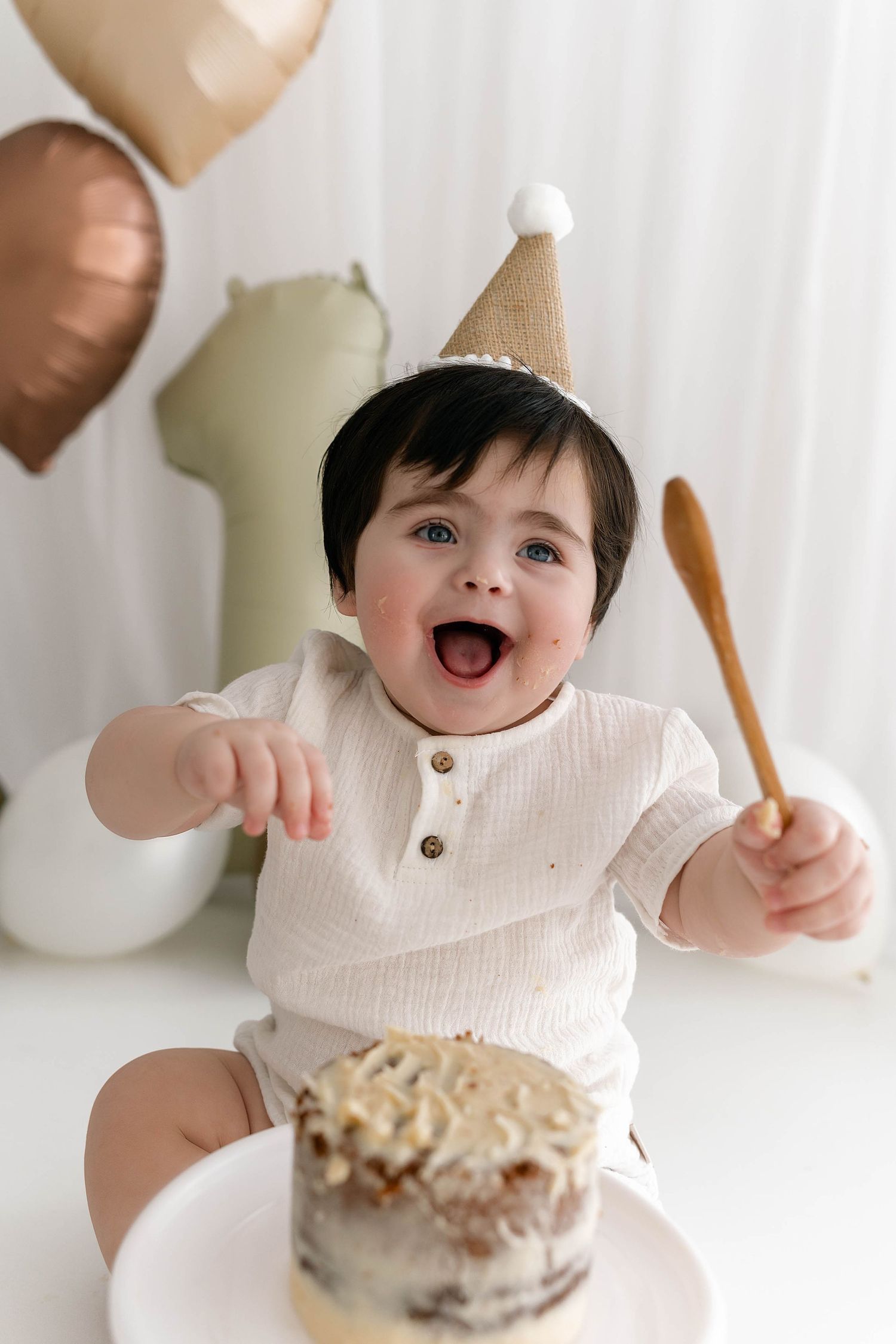 Small child in party hat sits excitedly at table with cake and wooden spoon during birthday celebration.