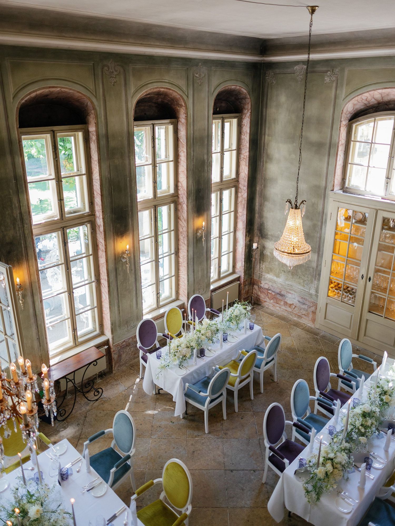 Elegant wedding reception setup in historic venue with sunlit arched windows, white tablecloths and pastel blue chairs.