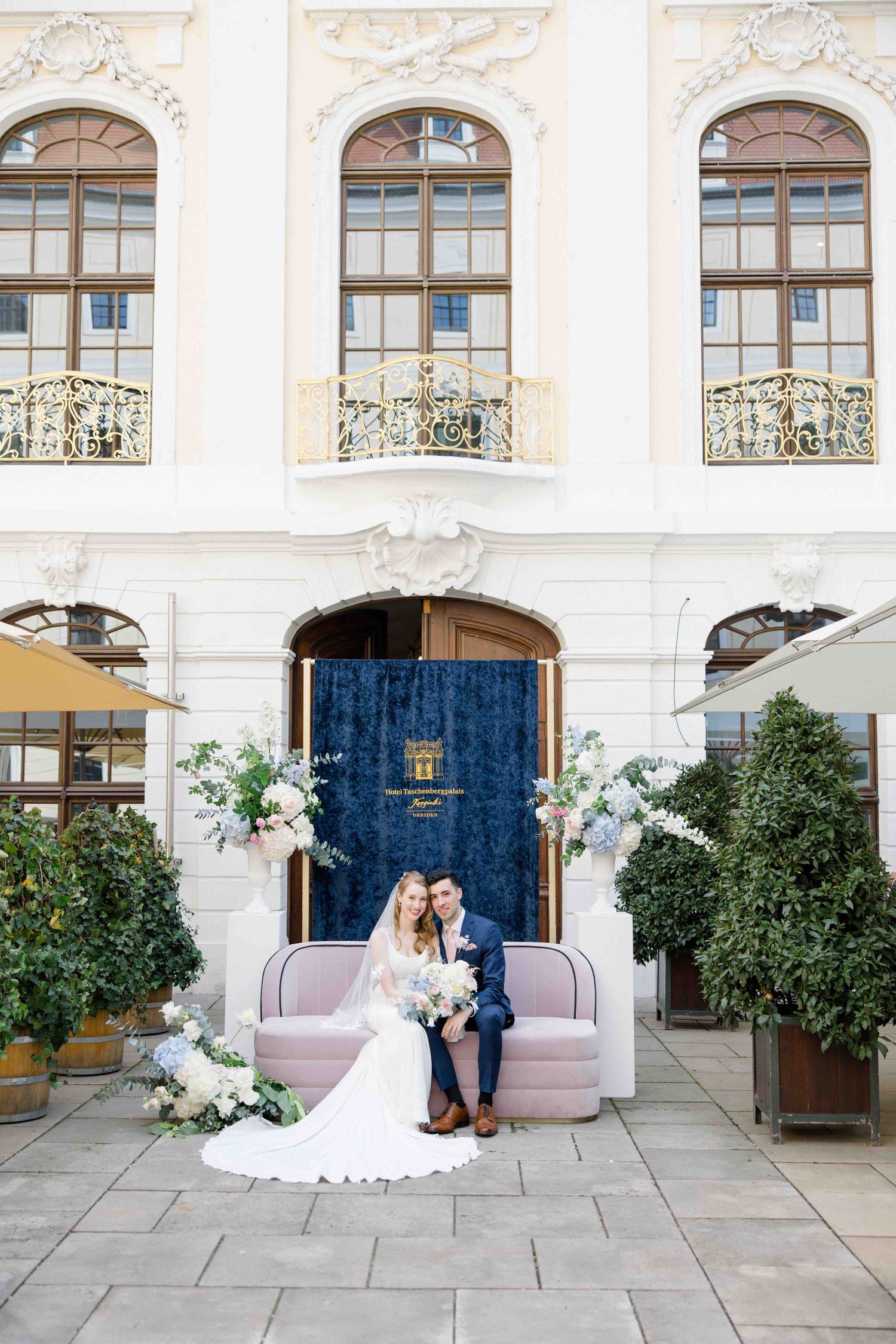 A romantic wedding scene set against an elegant white building with arched windows and classical architecture details.
