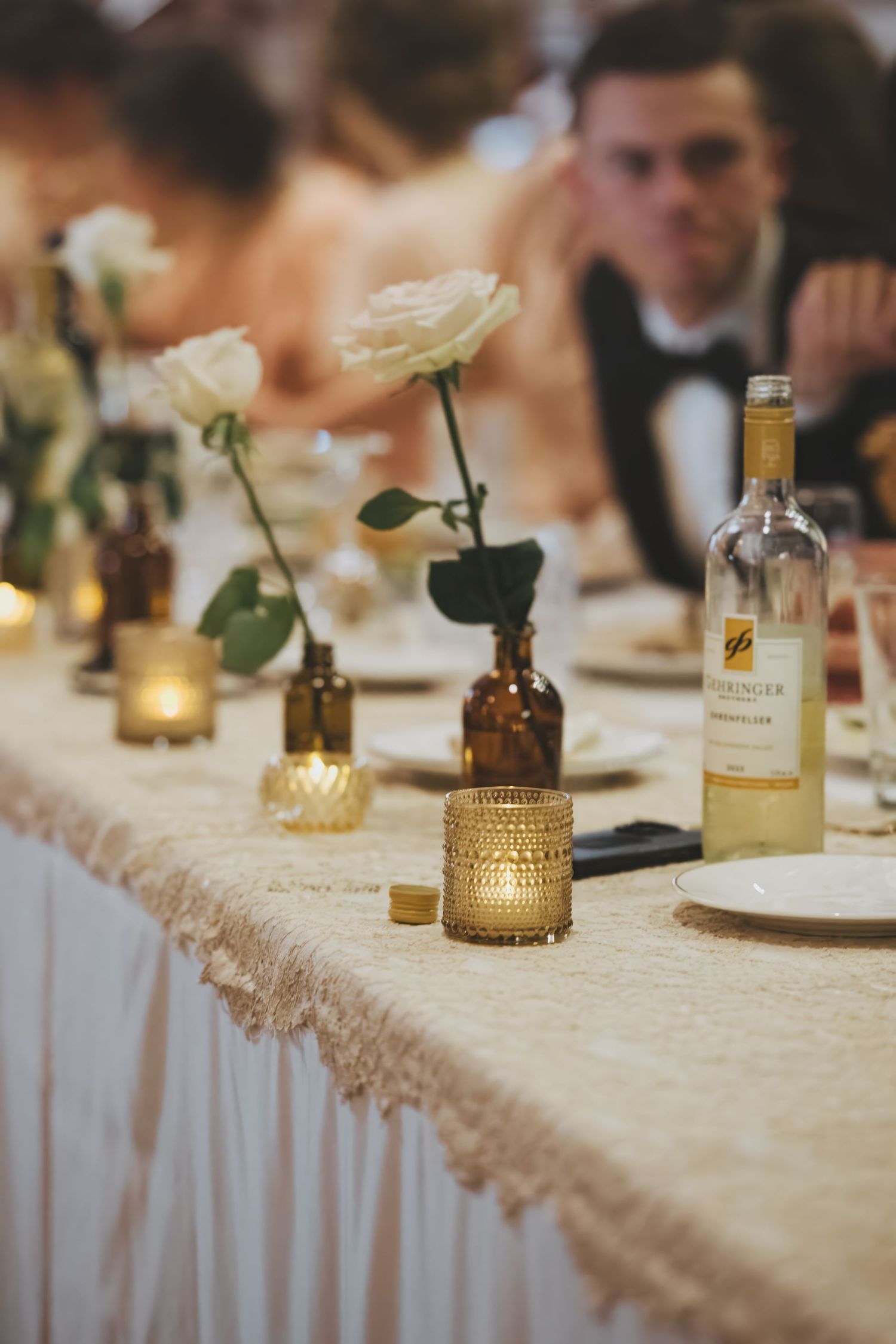 Wedding table decor featuring delicate white roses in amber glass vases, candlelight, and a bottle of wine on lace tablecloth.