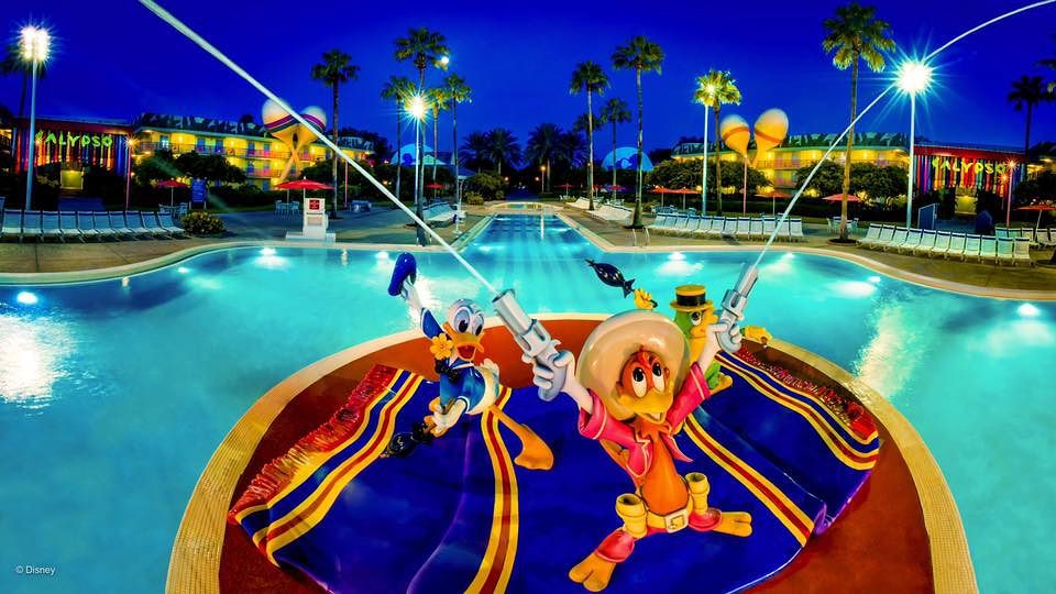 Illuminated pool at night featuring Disney character statues and palm trees at All-Star Resort.