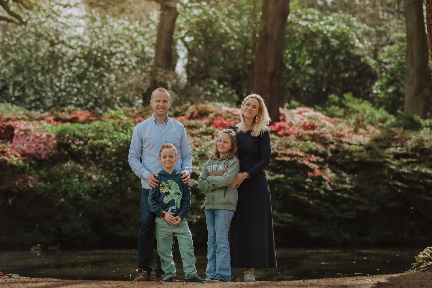 A family group stands together outdoors in a garden setting with blooming azalea bushes and trees in soft sunlight.