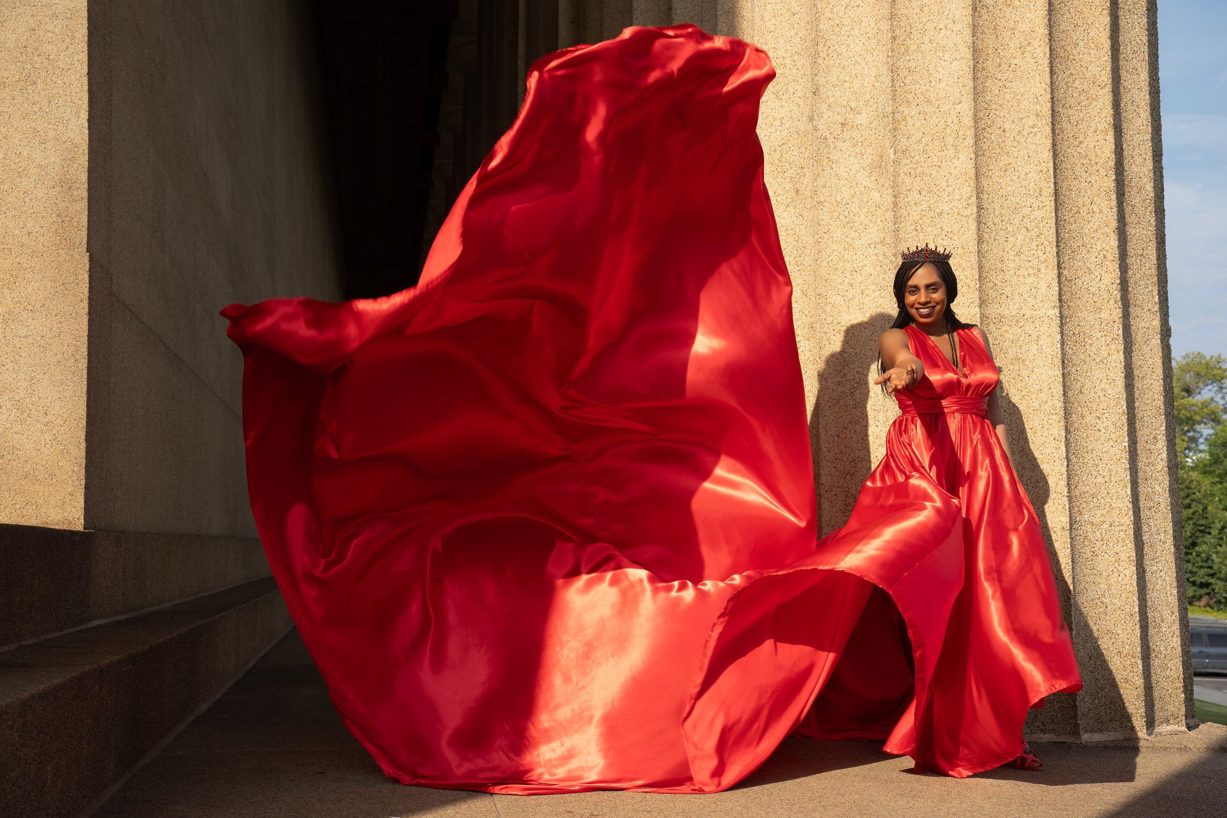 Red flowing fabric billows dramatically against a beige stone column backdrop in dramatic sunlight.
