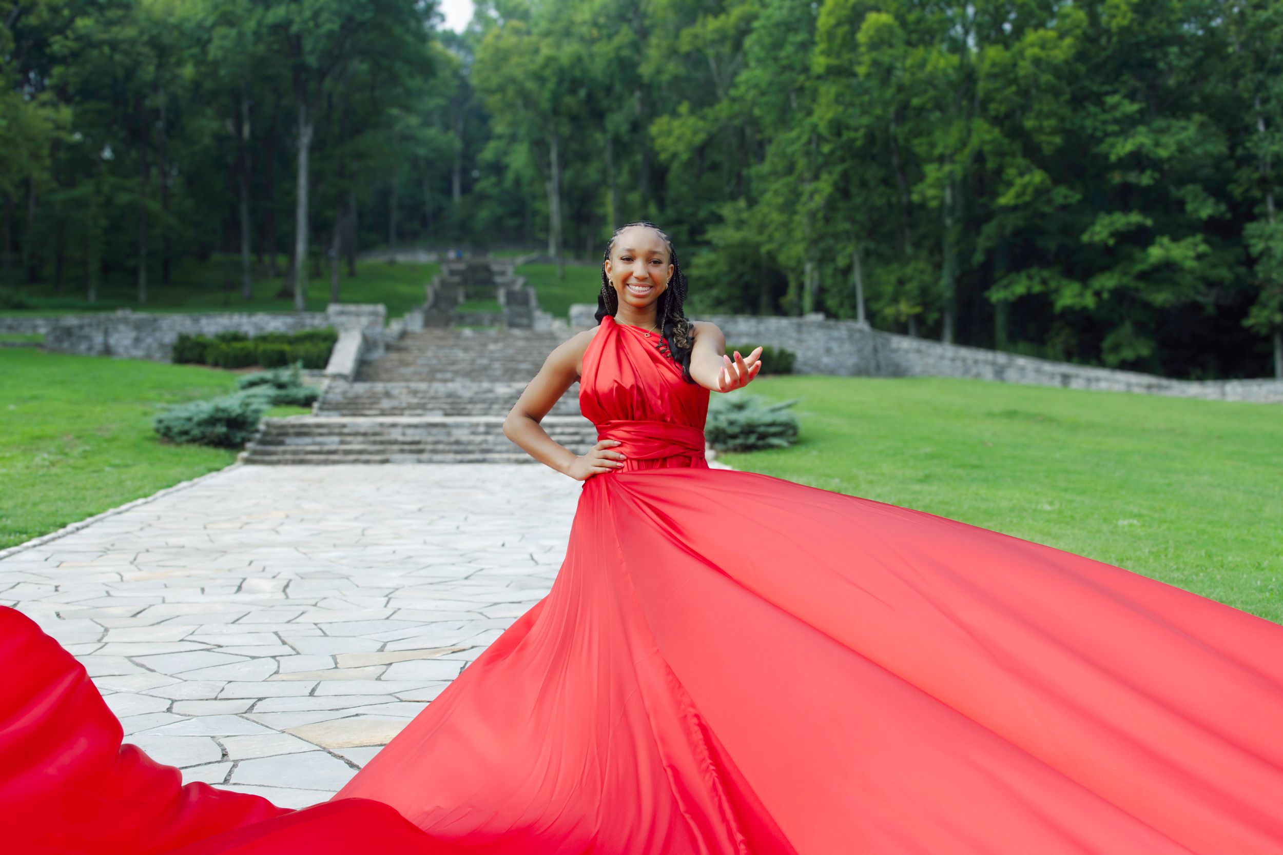 A flowing red gown with a dramatic train creates a striking contrast against a green garden landscape with stone pathways.