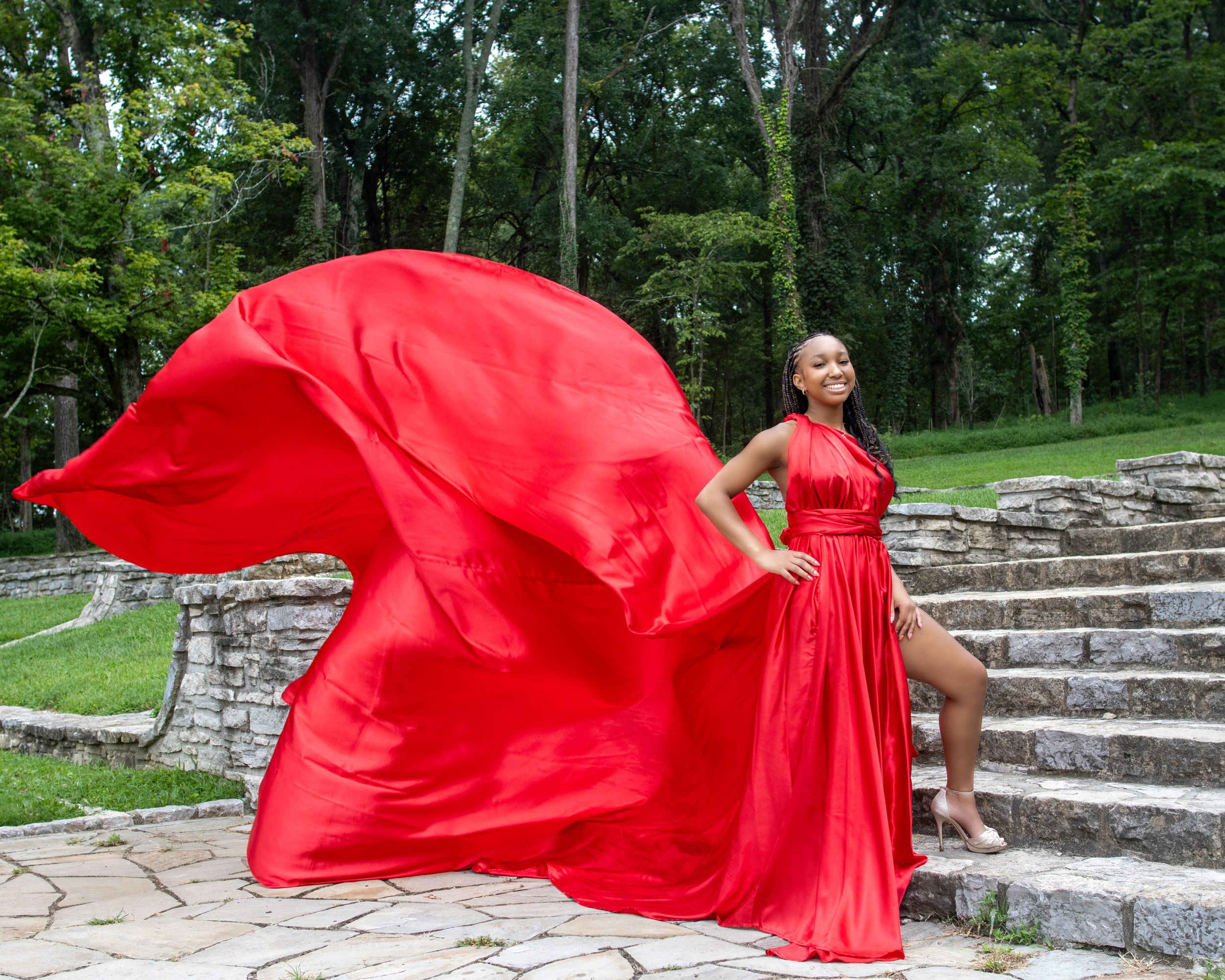 A flowing red evening gown with dramatic billowing fabric photographed on stone steps in a garden setting.