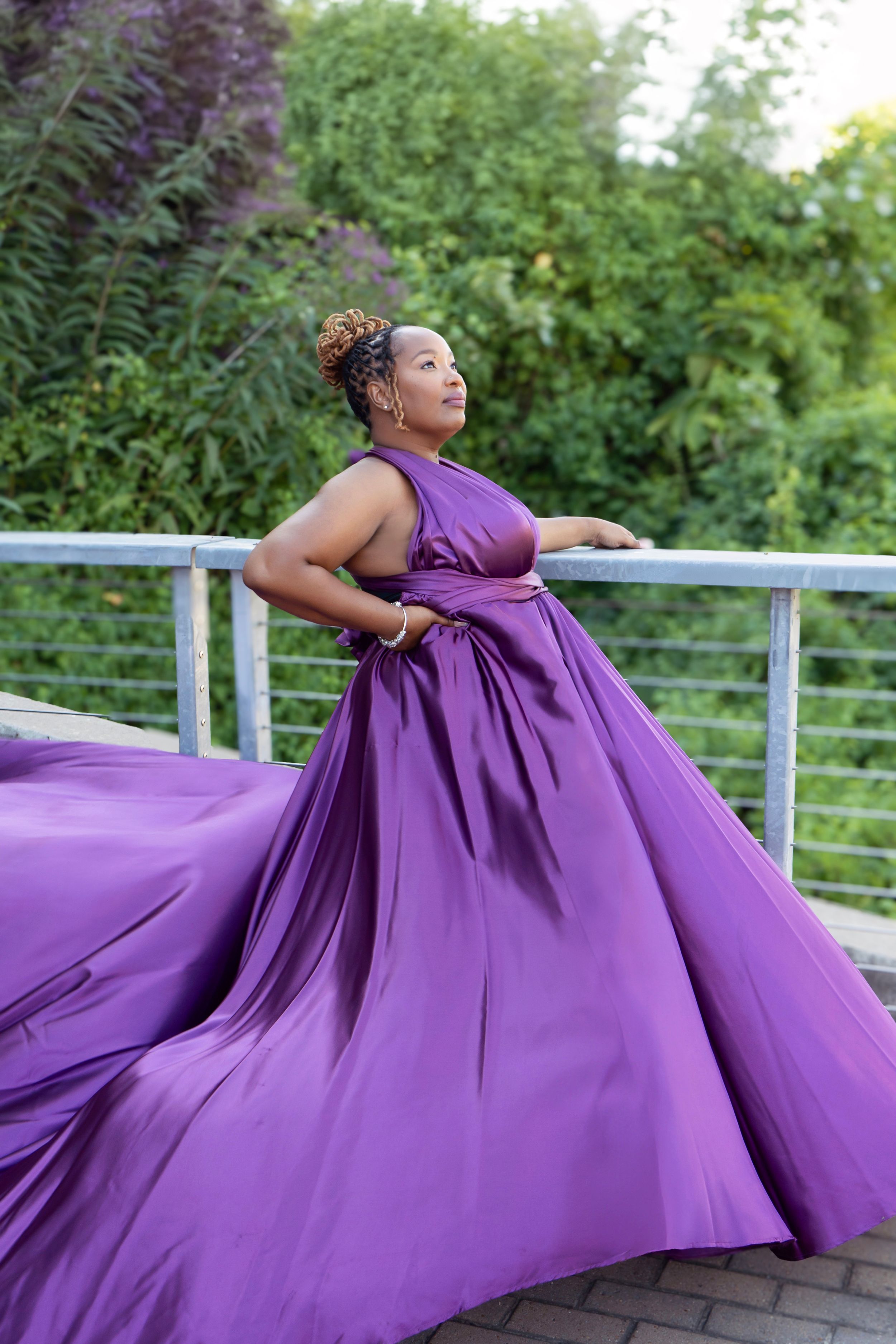 A flowing purple ball gown with dramatic movement swirls on an outdoor balcony against green foliage.