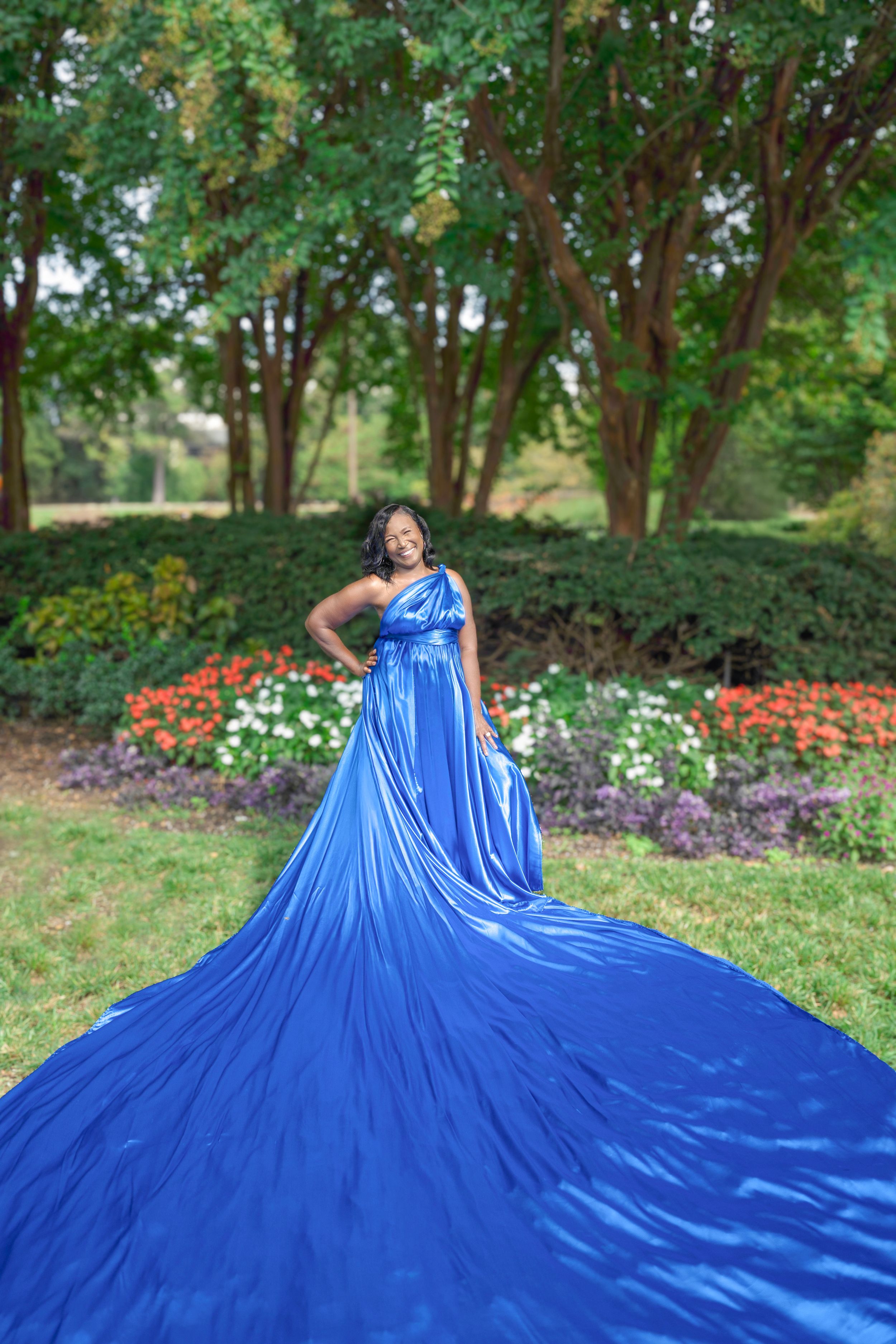 A sweeping floor-length royal blue ball gown flows dramatically across a garden setting with blooming flowers.