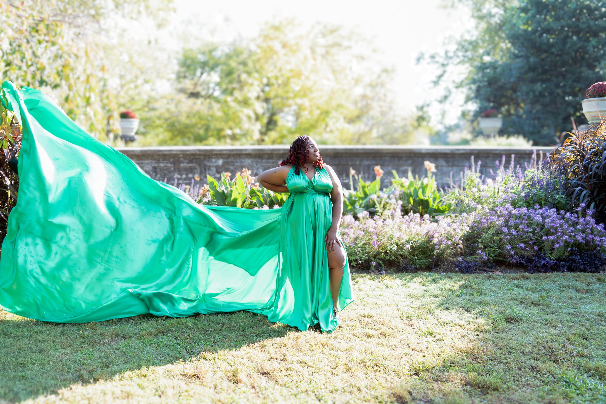 Woman in a flowing emerald green gown poses elegantly in a sunny garden setting with billowing fabric movement.
