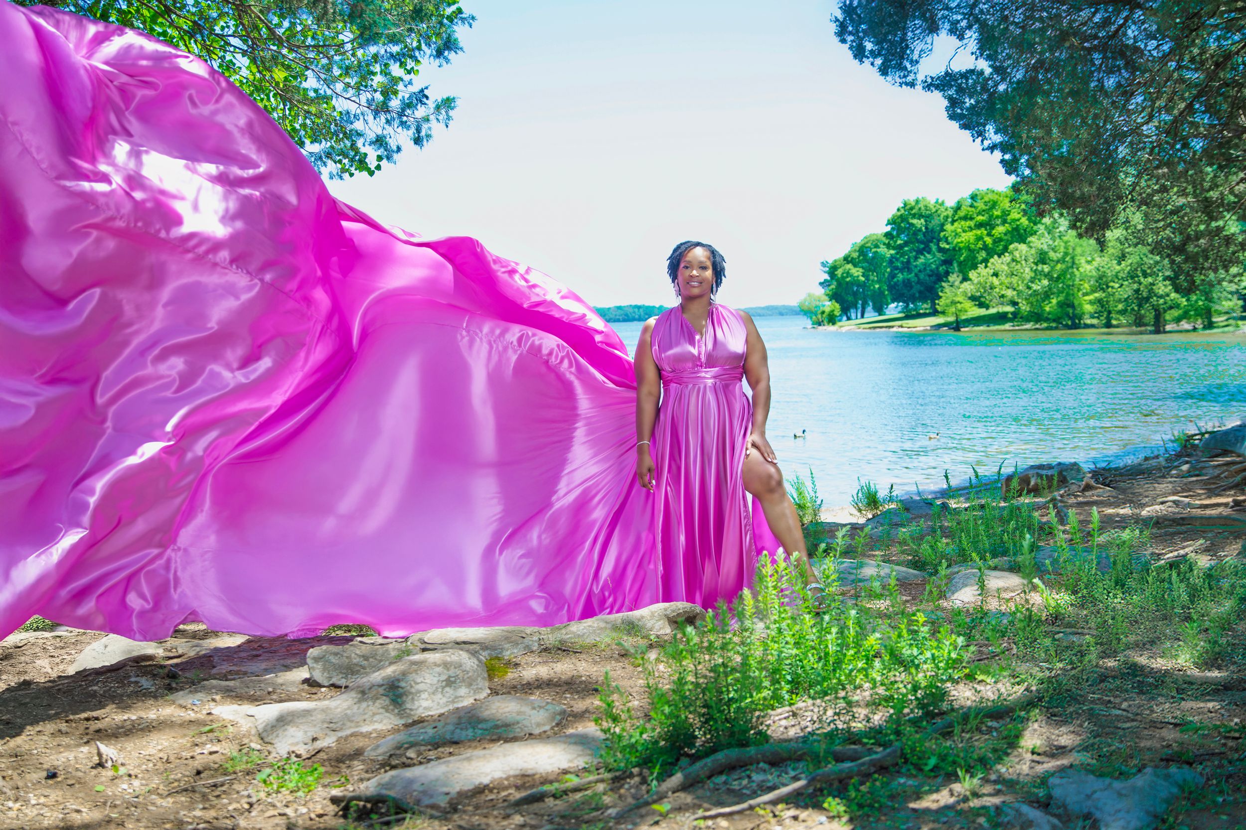 A flowing pink dress billows in the wind against a scenic lakeside backdrop on a sunny summer day.